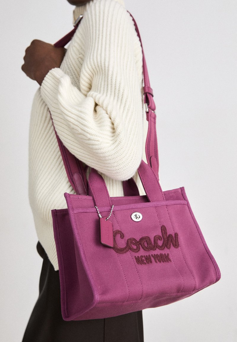 Purple canvas tote bag with embossed "Coach New York" logo, double handles, and a detachable tag, worn on a person in a knit sweater.