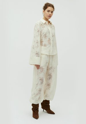 Kobieta ubrana w prześwitującą białą bluzkę z kwiatowej koronki i dopasowane szerokie spodnie, zestawione z brązowymi luźnymi kozaczkami na wysokim obcasie na białym tle.