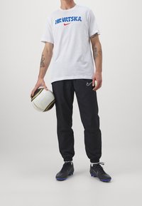 Valkoinen Nike t-paita, jossa on sininen ja punainen "HRVATSKA"-teksti, mustat Nike-harrastusshortsit ja mustat jalkapallokengät, jotka pitävät käsissään teksturoidun jalkapallon.