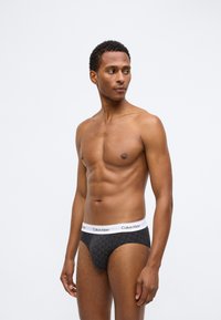 Čierne slipy s geometrickým vzorom, s bielym pásom Calvin Klein. Materiál vyzerá hladko a elasticky s tesným strihom.