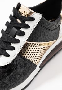 Black and white sneaker with black laces, gold metallic and patterned accents, textured side panel, and Michael Kors branding on the tongue and side.
