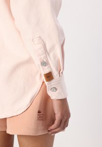 Svetlo roza majica z dolgimi rokavi in gumbi na manšetah, ki je v paru z usnjenim logotipom v bež barvi, v kombinaciji z ujemajočimi se roza kratkimi hlačami z natisnjenim blagovno znamko.