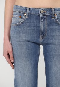Jean en denim bleu moyen avec bouton sur le devant et poches, porté par une personne avec la main gauche détendue le long de la cuisse.
