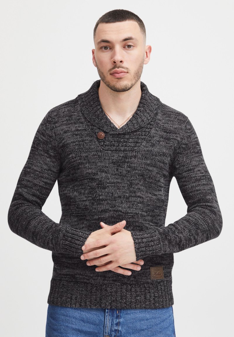 Gestrickter schwarzer und grauer Pullover mit Schalkragen, Holzknopf-Detail, strukturiertem Stoff und gerippten Bündchen, kombiniert mit Jeans.