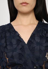 Kék blúz virágos csipke dizájnnal, V nyakkivágással és rövid ujjal. A frontrészen texturált virágminták és fekete gombok díszítik.