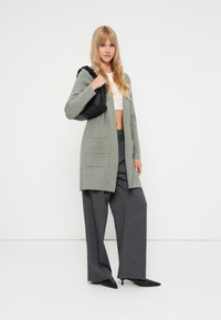 Πράσινο μακριά cardigan με δύο μπροστινές τσέπες, φορεμένο πάνω από ένα λευκό κοντομάνικο τοπ. Συνδυασμένο με γκρι φαρδιά παντελόνια και μαύρες μυτερές γόβες.