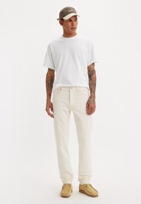 Lichtgekleurde jeans, wit T-shirt en beige schoenen. Grijze pet met merklogo. Kenmerken zijn korte mouwen en een zachte textuur van de stof.