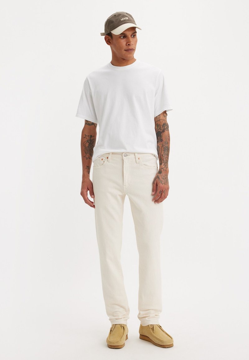 Lichtgekleurde jeans, wit T-shirt en beige schoenen. Grijze pet met merklogo. Kenmerken zijn korte mouwen en een zachte textuur van de stof.