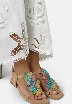 Pies con sandalias beige decoradas con hojas de cuentas multicolores, combinadas con tela blanca que presenta patrones florales bordados con recortes.