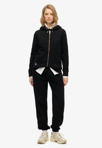 Sudadera negra con cremallera y cordón, bolsillos frontales y logo bordado, combinada con joggers negros y zapatillas crema. Textura suave.