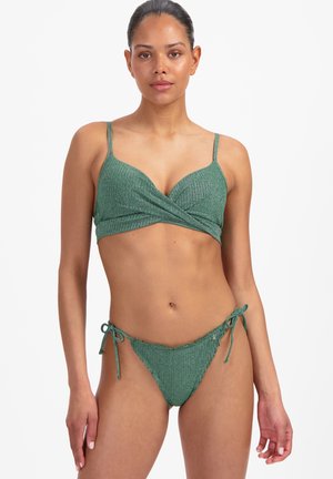 Femme portant un bikini vert texturé avec un haut croisé et un bas à nouer sur les côtés, debout devant un fond blanc uni.