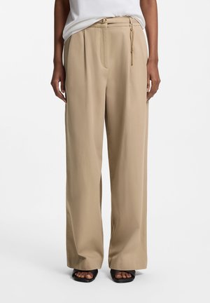 Person iført beige bukser med vide ben, en hvid top og sorte åbne sko, stående mod en ensartet hvid baggrund.