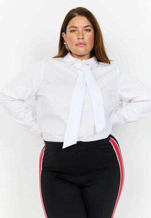 Chemise blanche à col montant avec un détail de nœud papillon, associée à des leggings noirs avec des rayures latérales rouges et blanches.