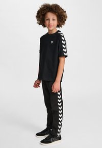 T-shirt nera con strisce a zigzag bianche sulle maniche, caratterizzata da un piccolo logo. Abbinata a pantaloni neri con accenti laterali a zigzag coordinati.