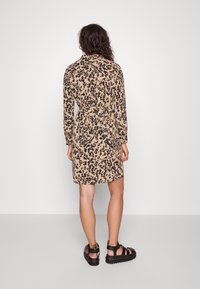 Tělově hnědé šaty s černým leopardím potiskem, s dlouhými rukávy, límečkem a páskem v pase. Nošeny s černými sandály s pásky.