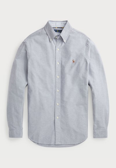 Chemise à manches longues bleu clair, boutonnée, en tissu tissé. Elle possède un col pointu, un logo sur la poitrine et des boutons blancs.