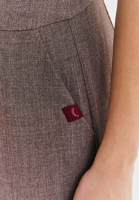 Detail vrecka odevu z hnedej textúrovanej látky s malou bordovou značkou so symbolom pripomínajúcim biele mihalnice.