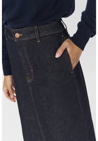 Personne portant une jupe en denim bleu foncé avec la main dans la poche latérale et un pull à manches longues bleu marine visible à la taille et aux manches.