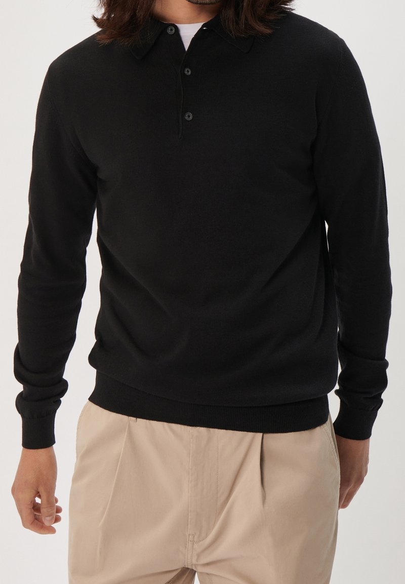 Homme portant un polo noir à manches longues et un pantalon beige à plis, se tenant devant un fond uni.