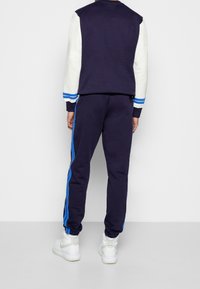 Námornická tepláková souprava s jednobarevným vrškem, bílými rukávy a modrými pruhy, doplněná o odpovídající kalhoty s bočními modrými pruhy a manžetami na kotnících.