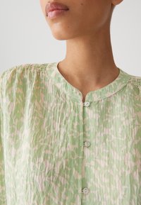 Blusa verde chiaro con un motivo astratto in rosa pallido. Presenta un collo rotondo, chiusure a bottone e un tessuto testurizzato con delicate pince.