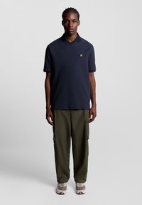 Marinblå polo med en strukturerad diamantmönster och en gul logotyp, matchad med olivgröna cargobyxor och ljusa sneakers.