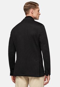 Black textured blazer with a notched collar, single vent, and two side pockets. Tailored fit, worn over light-colored trousers.