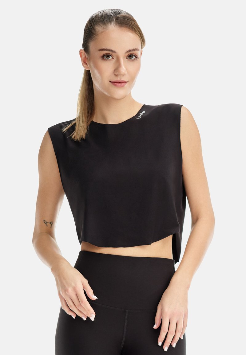 Junge Frau mit hellbraunen Haaren im Pferdeschwanz, die ein schwarzes ärmelloses Cropped-Top und hoch taillierte schwarze Leggings trägt.