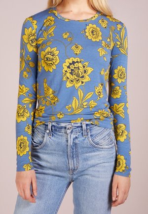 Bluse mit langen Ärmeln in Blau, verziert mit gelben Blumenmustern. Der Baumwollstoff hat eine glatte Textur, einen runden Halsausschnitt und eine figurbetonte Passform. Kombiniert mit hochgeschnittenen Jeans.