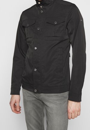 Homme portant une veste noire boutonnée avec des poches à rabat sur la poitrine et un pantalon gris clair, devant un fond uni.