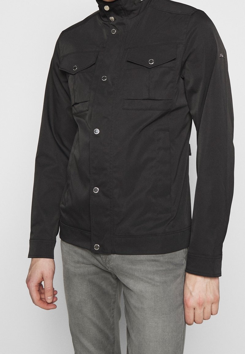 Homme portant une veste noire boutonnée avec des poches à rabat sur la poitrine et un pantalon gris clair, devant un fond uni.
