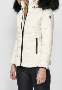 Giacca bianca gonfia con cappuccio in pelliccia sintetica nera, chiusura a zip e tasche laterali. Presenta una texture trapuntata e dettagli in metallo nero.