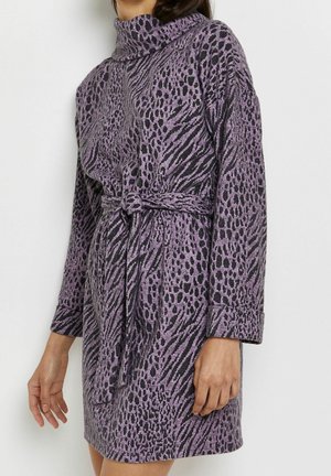 Nainen, joka pukeutunut vyölliseen, pitkähihainen violettiin mekkoon, jossa on musta eläinprintti ja korkea kaulus, seisoo yksinkertaisen valkoisen taustan edessä.