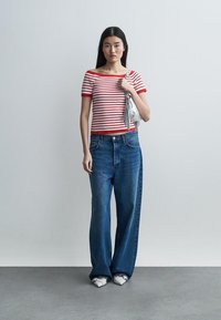 Γυναίκα που στέκεται, φορώντας μπλούζα με κόκκινες και λευκές ρίγες, φαρδιά μπλε τζιν παντελόνια, λευκά μυτερά παπούτσια και κρατώντας ασημένια τσάντα στον ώμο της.