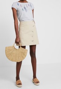 Camiseta gris con estampados de plumas, falda mini beige con botones, bolsa circular de paja tejida y sandalias de esparto color marrón.