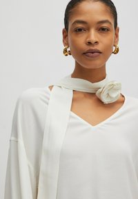 Weiße Bluse mit V-Ausschnitt, übergroßen Ärmeln und einem gebundenen Stoffakzent in Blumenform am Hals, kombiniert mit großen goldenen Creolen.