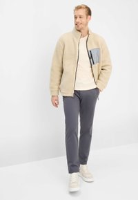 Fleecevest in beige met een grijs borstzakje, ritssluiting en hoge kraag; gecombineerd met grijze broek en lichte sneakers.