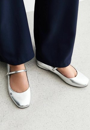 Ballerine argento con punta arrotondata e cinturino alla caviglia, caratterizzate da una superficie metallica liscia, indossate con pantaloni blu navy su una superficie chiara.