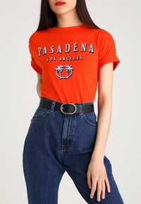 T-shirt en coton rouge avec "PASADENA LOS ANGELES" imprimé en blanc et bleu, associé à un jean en denim bleu taille haute et une ceinture noire.