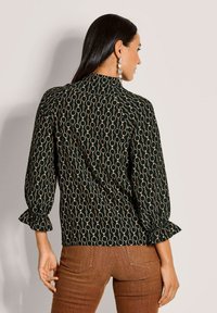 Schwarze Bluse mit einem goldenen, kreisförmigen Muster, langen Ärmeln mit Rüschensäumen und einem hohen Kragen. Das Modell trägt dazu braune Hosen.