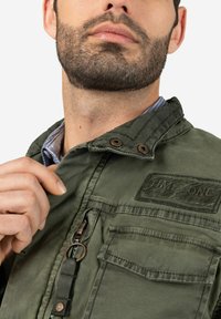 Olivgrüne Jacke mit Kragen, Reißverschluss, Brusttasche und einem Stoffetikett. Strukturiertes Material mit auffälligen Nähten und bronzenen Hardware-Akzenten.