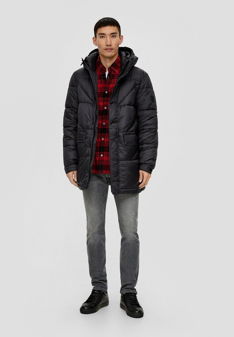 Parka negra acolchada con capucha, con bolsillos frontales y un cuello alto, llevada sobre una camisa de cuadros roja y jeans grises.