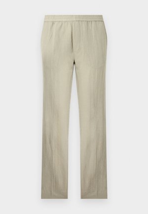 Homme aux cheveux bouclés portant une chemise grise à manches longues, un pantalon beige et des sandales marron foncé, debout avec une main dans la poche.
