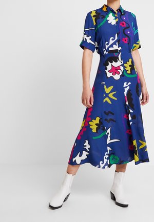 Kobieta w kolorowej, niebieskiej sukience midi z abstrakcyjnym kwiatowym wzorem i białych botkach, stojąca na tle jednolitego, jasnego tła.