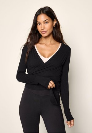 Giovane donna con lunghi capelli scuri indossa un top nero a incrocio sopra una canotta beige chiaro e pantaloni neri attillati su uno sfondo neutro.