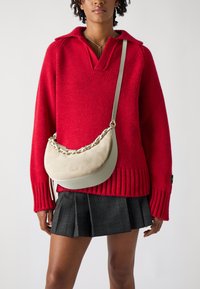 Rød strikket sweater med krave, parret med en plisseret sort ternet nederdel. Lys grå skuldertaske med en tekstureret overflade og kædedetalje.