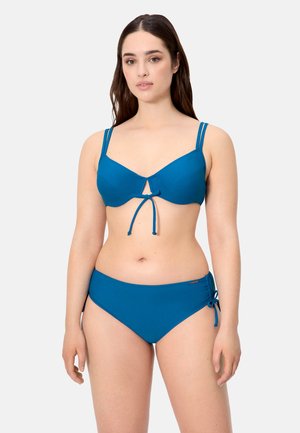 Mujer con bikini azul con tirantes dobles en los hombros y lazos ajustables en el centro superior y en la cadera izquierda, de pie sobre un fondo liso.