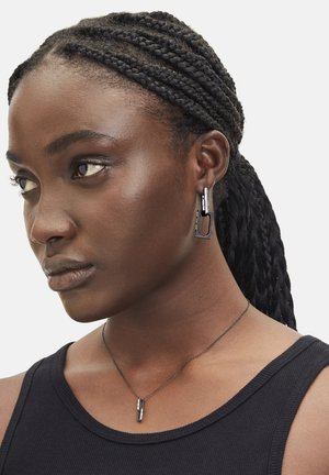 Schwarze Metallohrringe mit geometrischem Design und strukturierten Akzenten, kombiniert mit einer passenden, eleganten Halskette, getragen von einer Person in einem schwarzen Tanktop.