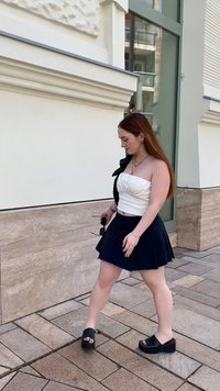 Top branco sem alças com textura franzida, saia mini plissada azul-marinho, sandálias de plataforma pretas e óculos de sol na mão; cabelo longo e liso.