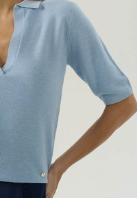 Svetlomodrý pletený top s krátkym rukávom a výstrihom do V, ktorý má osoba so založenou jednou rukou na páse na pozadí bez vzoru.